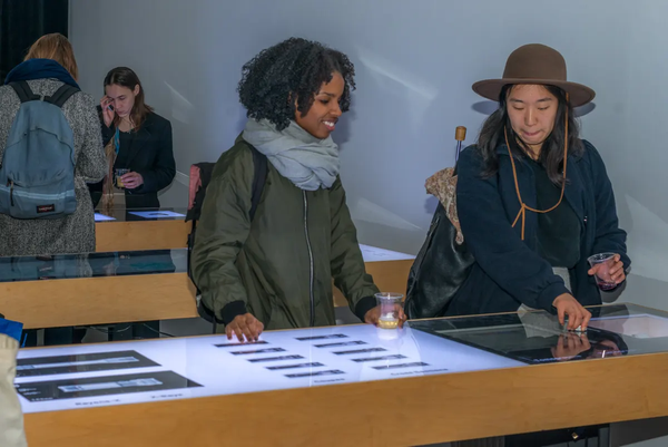 Dans une galerie, deux personnes interagissent avec un module. Un personne semble regarder une autre personne alors qu'elle interagis avec l'interface. Cette personne utilise l'interface entre l'écran et les deux vraies lampes de poche. 