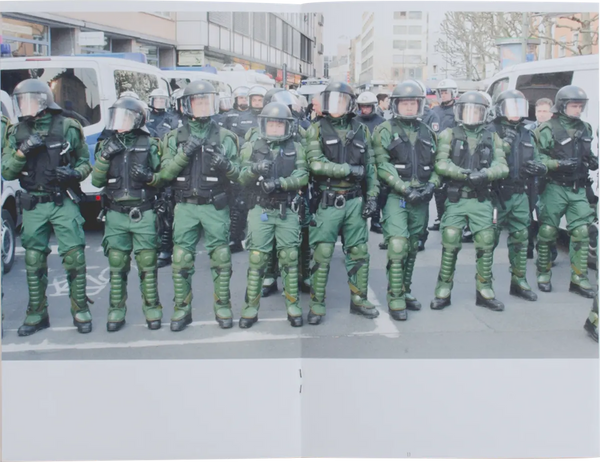 Open book. A photo covers both pages. The photo shows a row of police in green uniforms blocking a street. 
