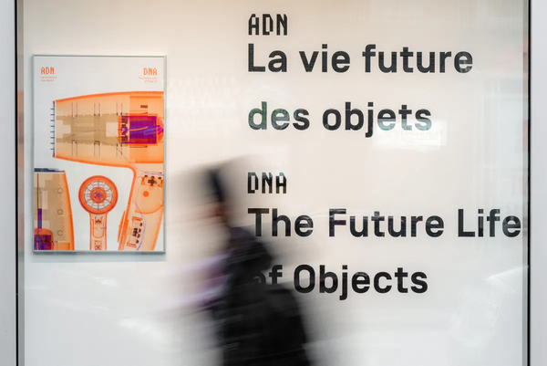 Affiche et nom de l'exposition derrière ue vitrine passante. On y lit ADN, la vie future des objets en français et en anglais. Le flou d'une personne passe devant la vitrine rapidement.