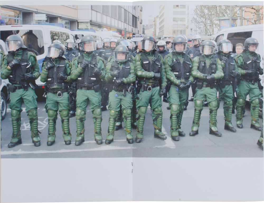 Livre ouvert. Une photo couvre les deux pages. Dans la photo, une rangée de policiers en habit kaki bloque l'accès à une rue.